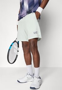 Helle mintgrüne Sportshorts mit Taschen, seitlichen Schlitzen und EA7-Logo. Getragen mit weißen Sportschuhen und passenden Socken in Crew-Länge. Tennisschläger gehalten.