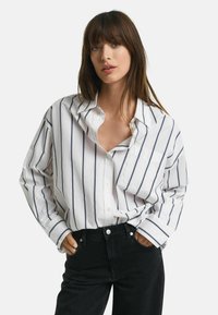 Witte katoenen blouse met marineblauwe verticale strepen, een losse pasvorm, knoopsluiting aan de voorkant, een borstzak en opgerolde mouwen, gecombineerd met zwarte jeans.