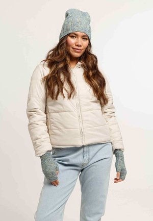 Beige quilted jacket with a front zipper, paired with a light blue knitted beanie and fingerless gloves. Blue high-waisted jeans complete the outfit.