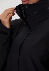 Černá voděodolná bunda s vysokým límcem, zipem a patenty. Materiál vypadá hladce s jemným leskem a texturovanými akcenty.