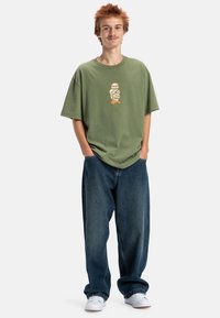 Olivegrynowy, oversize T-shirt z nadrukiem graficznym, zestawiony z luźnymi, niebieskimi dżinsami i białymi sneakersami. Swobodny outfit o luźnym kroju.