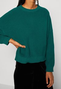 Miętowy sweter na drutach o luźnym kroju, szerokim dekolcie i teksturowanym wzorze. Posiada ściągacze na rękawach i krótki dół, stylizowany z czarnymi aksamitnymi spodniami.