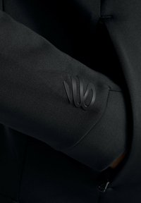 Black blazer sleeve with a smooth texture, featuring a prominent black logo accent on the cuff. Tailored fit with subtle detailing.