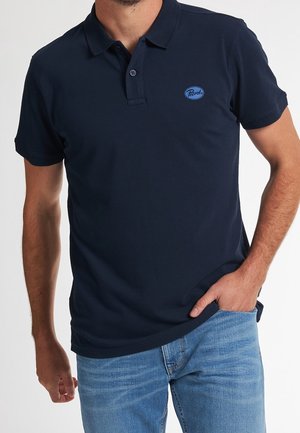 Mand iført en mørkeblå kortærmet polo med en lille blå logo på brystet og lyseblå jeans, hånd i lommen.