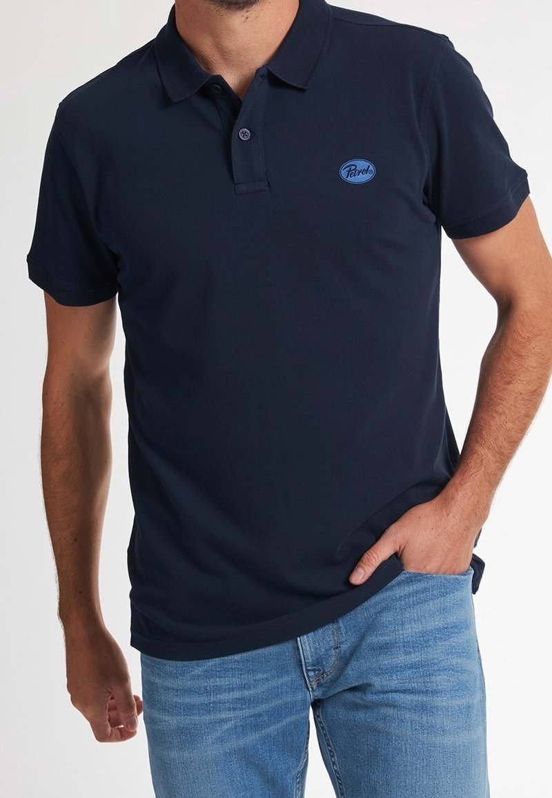 Homme portant un polo bleu marine à manches courtes avec un petit logo bleu sur la poitrine et un jean bleu clair, main dans la poche.