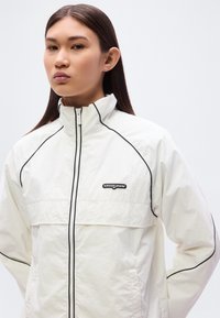 Λευκό μπουφάν με φερμουάρ και μαύρες λεπτομέρειες, με λογότυπο στο στήθος. Διαθέτει ψηλό γιακά και άνετη εφαρμογή. Λείο, ελαφρύ ύφασμα.