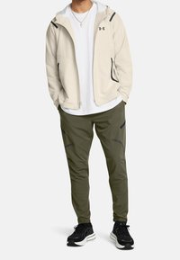 Ljusbeige zip-up jacka med svarta detaljer, i kombination med olivgröna byxor med dragkedjefickor och stiliga svarta sneakers.