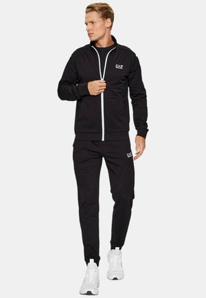 Tuta sportiva nera con giacca a zip e pantaloni abbinati. Presenta dettagli bianchi e logo, realizzata in un materiale liscio e flessibile.