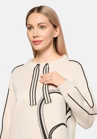 Jasnobeżowa bluza z okrągłym dekoltem, czarnymi liniami akcentującymi oraz sznurowadłami w paski. Na przodzie duży czarny wzór w kształcie serca.