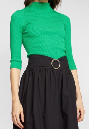 Femme portant un haut côtelé vert vif à manches trois-quarts et une jupe noire taille haute avec une boucle de ceinture circulaire argentée.