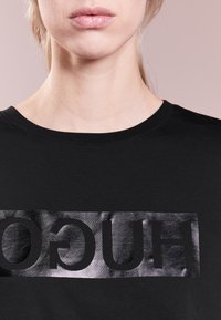 Czarna koszulka z połyskującym srebrnym logo "HUGO" na przodzie, okrągły dekolt i miękka tkanina.