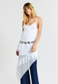 Donna che indossa un top asimmetrico bianco in pizzo con volant e una cintura argentata sopra pantaloni a gamba larga blu scuro su uno sfondo neutro.