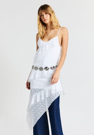 Donna che indossa un top asimmetrico bianco in pizzo con volant e una cintura argentata sopra pantaloni a gamba larga blu scuro su uno sfondo neutro.
