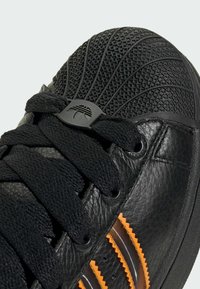 Baskets en cuir noir avec embout texturé, lacets noirs et accents rayés orange sur le côté.