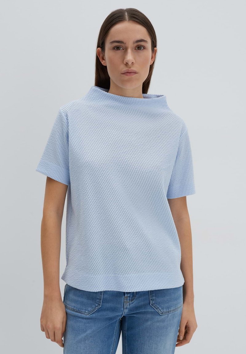 Himmelblause, kurzärmliges Oberteil mit hohem Ausschnitt, versehen mit einem strukturierten Muster. Kombiniert mit blauen Jeans, vor einem neutralen Hintergrund.