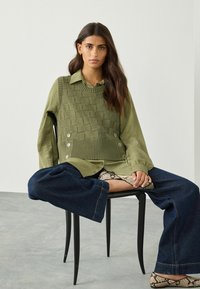 Olivgrön stickad väst över en långärmad olivgrön skjorta, i kombination med löst sittande mörka jeans. Påtagliga knappdetaljer och texturerade mönster.
