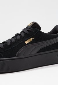 Sneaker in suede nera con una spessa suola piatta nera, lacci piatti neri e un logo Puma dorato sul lato e sulla linguetta.