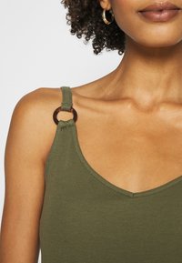 Olive green tank top with ribbed texture, featuring thin straps and a round, tortoiseshell accent on the shoulder.