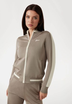 Bežna zip-up jakna z belimi črtami na rokavih in usklajenim pasom. Mehka tkanina z vezenim logotipom "Guess" na prsih.