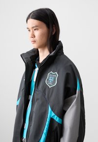 Black and gray windbreaker with turquoise accents, high collar, and shield logo featuring "BOTTER ANTWERP." Made from lightweight, smooth material.