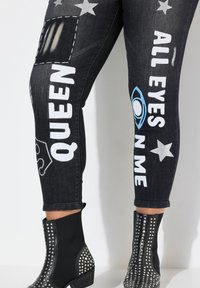 Jeans de mezclilla negra con estampados gráficos, que presentan texto blanco y patrones de estrellas. Combinados con botas negras hasta el tobillo adornadas con tachuelas de plata.