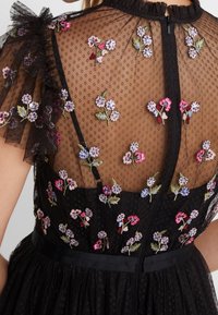Robe noire avec un dos en mesh transparent brodé de motifs floraux en rose et bleu, manches volantées, et taille cintrée avec un ruban.
