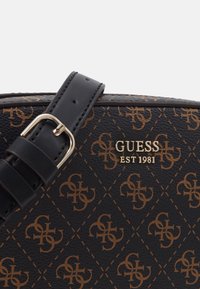 Borsa a mano sintetica nera con un motivo logo ripetuto marrone, decorazioni dorate e una tracolla in pelle nera con una fibbia metallica.