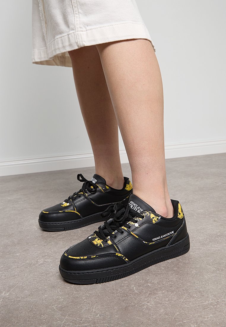 Las zapatillas negras de cuero presentan acentos gráficos amarillos, cordones planos y una suela de goma texturizada. La marca es visible en el lateral.