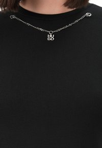 Silver chain necklace with "HUGO" pendant worn over a black ribbed fabric top with two metal eyelets near the collarbone.