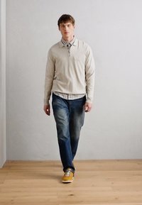 Beige pulóver, kockás ing fölött, kék farmernadrággal és sárga sportcipőkkel, egy sima fal és fa padló előtt.