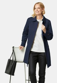 Marineblauer Trenchcoat aus wasserabweisendem Stoff, mit Knopfverschluss, tailliertem Schnitt und Seitentaschen. Schwarze Tragetasche auf einem Stuhl.