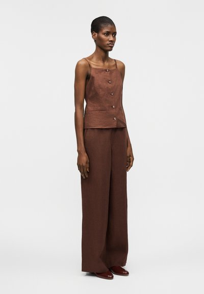 Γυναίκα που φοράει αμάνικο καφέ τοπ με κουμπιά και λεπτές τιράντες, ασορτί παντελόνι με φαρδιά μπατζάκια και καφέ παπούτσια, στέκεται μπροστά σε απλό φόντο.