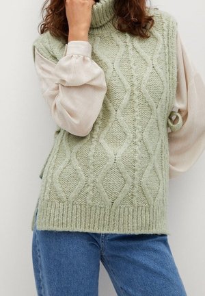 Frau trägt eine hellgrüne gestrickte ärmellose Weste über einer beigen Bluse mit langen Ärmeln und blauen Jeans, posiert mit der Hand am Gesicht.