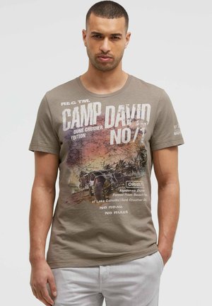 Man wearing brown graphic T-shirt with desert and off-road vehicle print, paired with light-colored pants, standing against plain background.