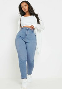 Siniset korkeavyötäröiset farkut, joissa on sileä rakenne, yhdistettynä valkoiseen lyhyeseen teeppariin ja kevyeseen valkoiseen neuletakkiin. Valkoiset lenkkarit.