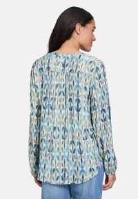 Femme avec de longs cheveux bruns portant un chemisier à manches longues bleu et beige à motifs et un jean bleu clair, de dos contre un fond blanc.