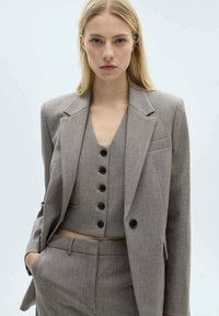 Costume gris cintré comprenant un blazer avec un col élancé, un gilet court avec des boutons noirs et un pantalon assorti à coupe droite.