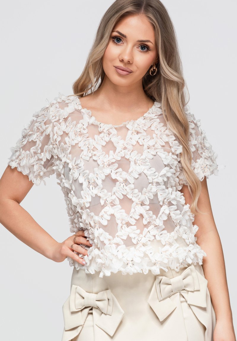 Femme aux longs cheveux blonds portant un haut blanc transparent à texture florale et un pantalon crème avec de grands nœuds à la taille.