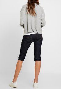 Suéter gris corto sobre una camiseta blanca y pantalones capri de mezclilla oscura. Zapatillas blancas completan el conjunto. Las características destacadas incluyen bolsillos traseros.