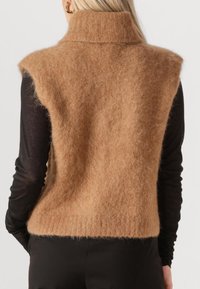 Blonde Person trägt eine flauschige braune ärmellose Rollkragenweste über einem langärmligen schwarzen Oberteil und schaut von der Kamera weg.