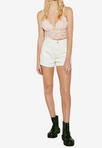 Krajkový bralet v jemně růžové, bílé džíny s krátkými nohavicemi a černé kotníčkové boty s robustními podrážkami a tkaničkami. Modelka stojí vzpřímeně s rukama na bocích.