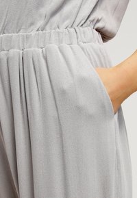 Jasnoszara tkanina o teksturowanym wykończeniu, z elastycznym paskiem w talii i bocznymi kieszeniami, prezentująca luźny, swobodny krój.