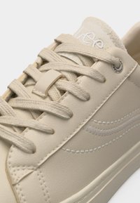 Κοντινό πλάνο μπεζ αθλητικού παπουτσιού με υφασμένα κορδόνια, κέντημα σε κυματιστό μοτίβο και διακοσμητική τρύπα κορδονιών με λογότυπο πάνω σε λείο δέρμα.