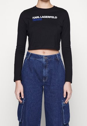 Personne portant un top court noir à manches longues avec le texte ""Karl Lagerfeld Jeans"" et un pantalon cargo en denim bleu taille haute avec des poches latérales.