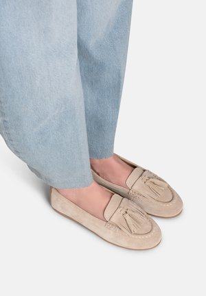 Mocassini beige in pelle scamosciata con nappe indossati con jeans denim azzurri chiari, primo piano di piedi e parti inferiori delle gambe su sfondo bianco.