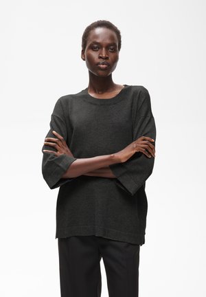 Femme aux cheveux courts portant un pull ample gris foncé et un pantalon noir, debout les bras croisés devant un fond blanc uni.