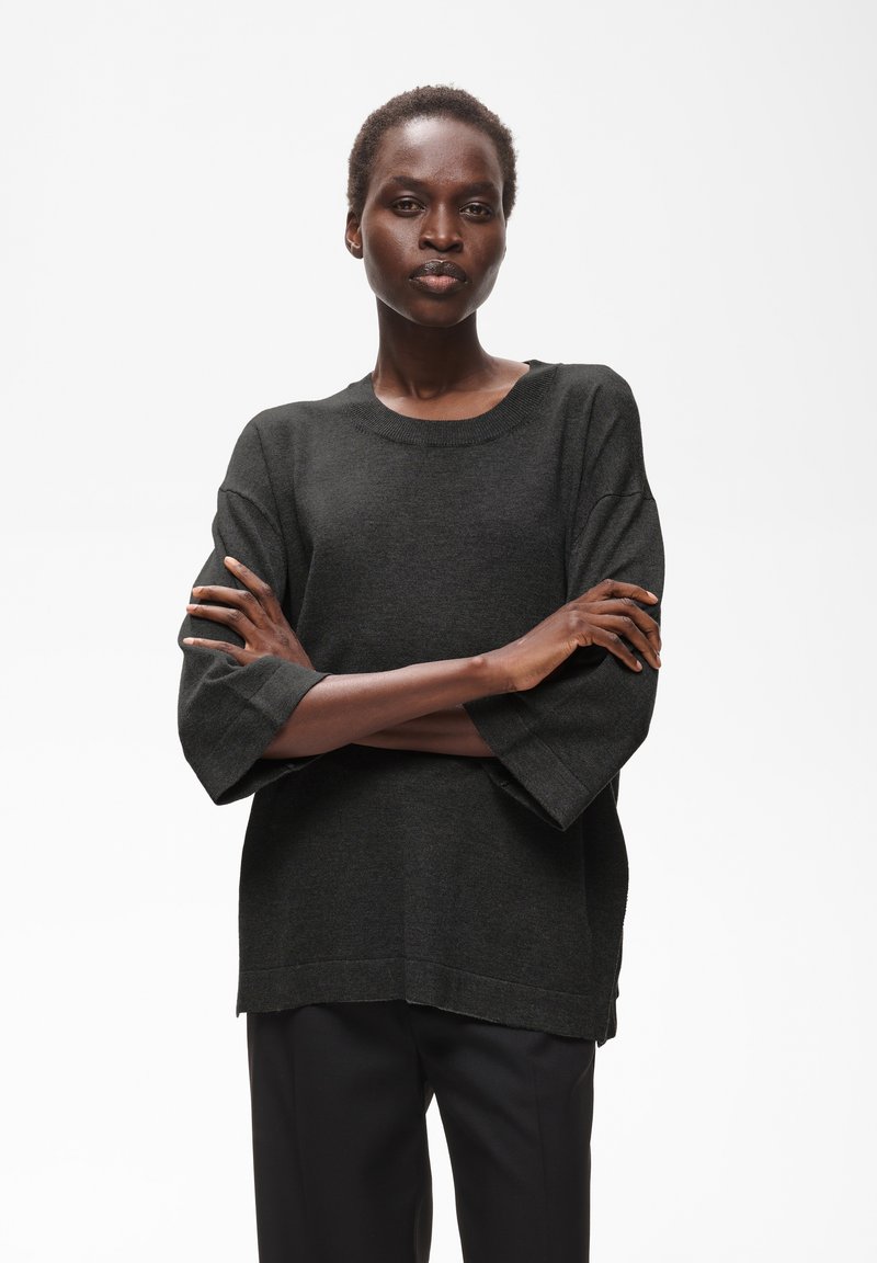 Femme aux cheveux courts portant un pull ample gris foncé et un pantalon noir, debout les bras croisés devant un fond blanc uni.
