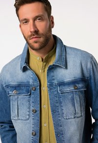 Chaqueta de mezclilla en un degradado de azul claro a oscuro, con botones de presión, dos bolsillos frontales, y combinada con una camisa de color verde claro con cuello.