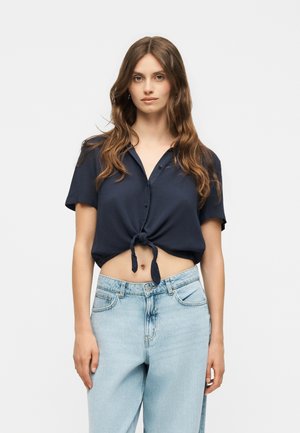 Femme aux longs cheveux ondulés portant une chemise bleu marine à manches courtes boutonnée et nouée, et un jean bleu clair taille haute, debout devant un fond uni.