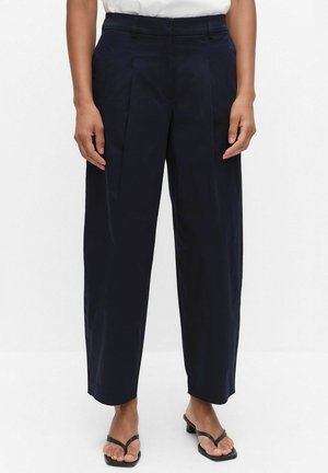 Persoon die marineblauwe wijde broek met voorplooien, witte top en zwarte sandalen met hak draagt, staand tegen een effen achtergrond.